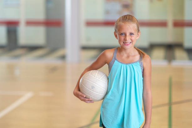 A young girl is standing in a school gym. She is holding a volleyball and smiling at the camera.
