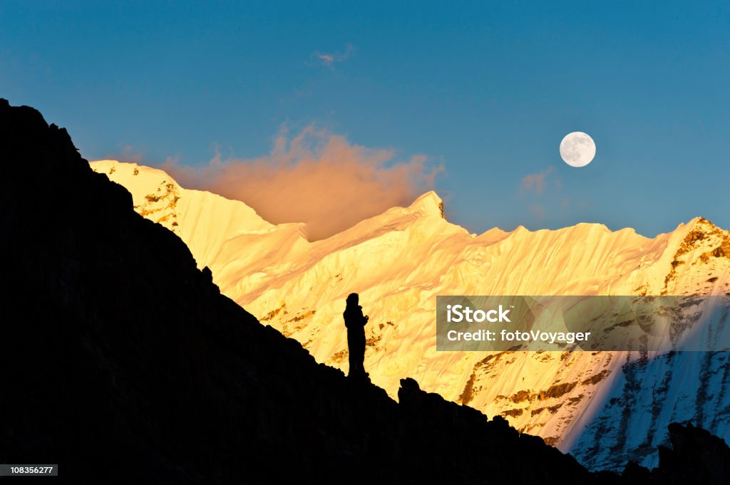 Mountaineer watching moon rise over golden sunset snow summit Himalayas  Adventure Stock Photo