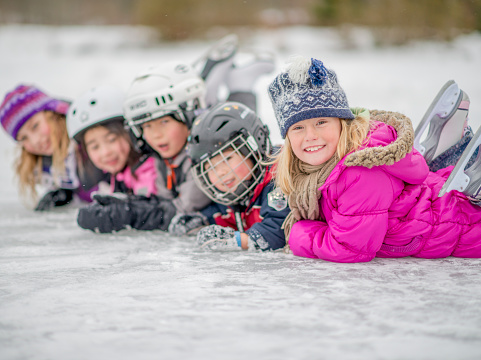 Five kids are lying on an outdoor ice rink in winter. They are wearing winter hats or skating helmets. The kids are smiling at the camera.