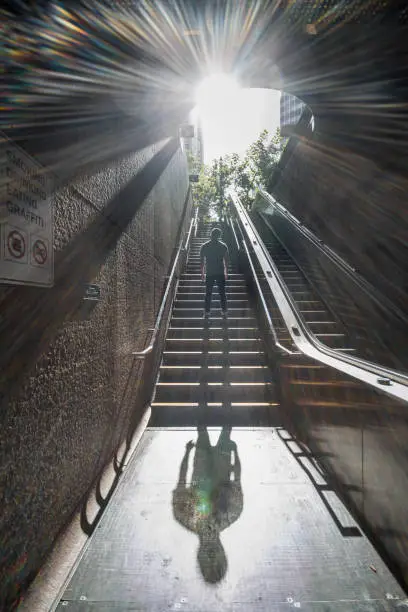 Photo of Bright sun beaming down on city fit man standing on subway stairs, large shadow casted.