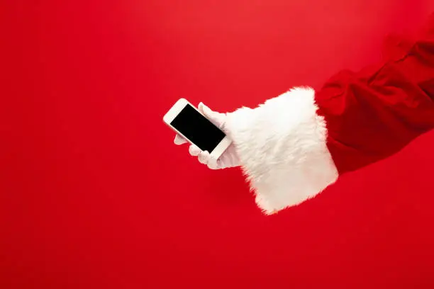 Santa Claus holding mobile phone ready for Christmas time on red studio background. The season, winter, holiday, celebration, gift concept