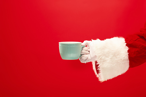 Christmas coffee. The Santa hand holding cup of cofee isolated on a red background with space for text. The season, winter, holiday, celebration, gift concept