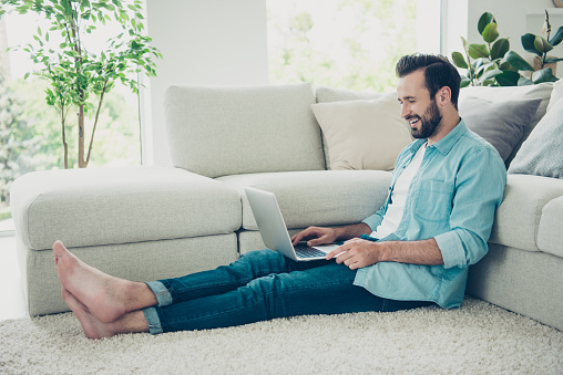 Full legs body size style stylish trendy cheerful attractive bearded man sit on comfort carpet floor in denim jeans blue shirt casual clothes inside bright light house interior using pc gadget