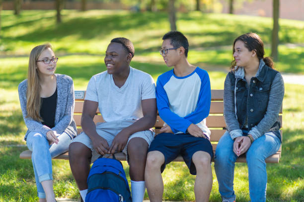 Teenagers hanging out at the park Four teenagers are sitting on a bench in a park and talking to each other. It is summertime. One boy has his backpack on the ground. teenagers only teenager multi ethnic group student stock pictures, royalty-free photos & images