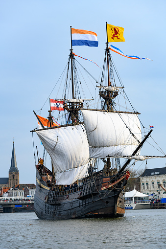 Old VOC sailing ship Halve Maen at the river IJssel during the 2018 Sail Kampen event in the Hanseatic league city of Kampen in Overijssel, The Netherlands. The Halve Maen was a trading ship of the Dutch East India Company (Dutch: Verenigde Oost-Indische Compagnie or VOC) and sailed into what is now New York Harbor in September 1609 during a search for a western passage to China. People on board are looking at the view and a crownd on the quay is watching the ships.