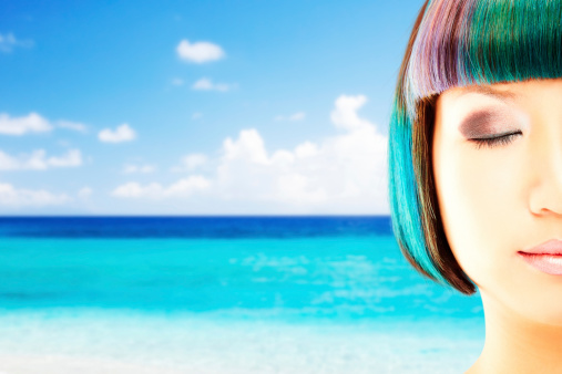 Cropped view of Asian woman with multi-colored hair and closed eyes in front of a tropical ocean background.