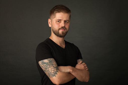 Studio portrait of an attractive 25 year old bearded tattooed man in a black t-shirt on a black background.