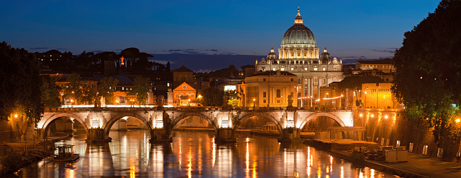 The iconic dome of the Basilica di San Pietro towering over the Vatican City and Bernini's magnificent marble angels on the Ponte Sant'Angelo reflecting in the River Tiber under panoramic sunset skies, Rome, Italy. ProPhoto RGB profile for maximum color fidelity and gamut.