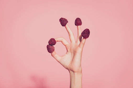 Close-up cropped nice well-groomed delicate hand five fingers sweet berries hats on nails showing ok-sign isolated over pink pastel background
