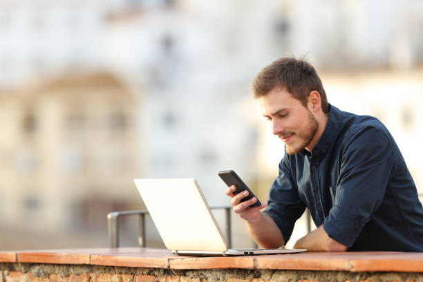 hombre con un ordenador portátil comprobando contenido del teléfono en un balcón - travel red vacations outdoors fotografías e imágenes de stock