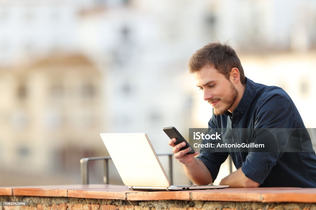 Man with a laptop checking phone content in a balcony Men Stock Photo