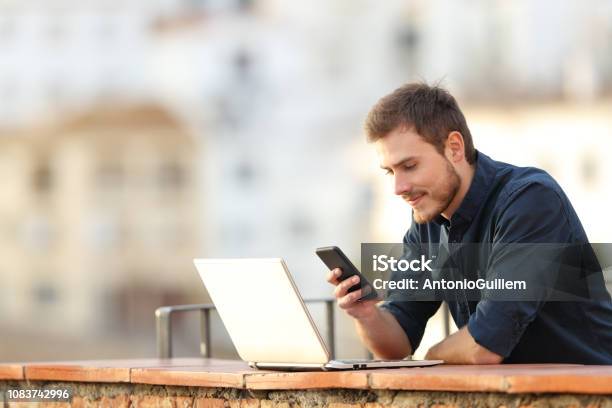 Man With A Laptop Checking Phone Content In A Balcony Stock Photo - Download Image Now