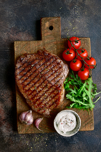 Roasted organic rancho beef steak with vegetables and garlic sauce on a wooden cutting board over dark slate, stone, metal or concrete background.Top view with copy space.