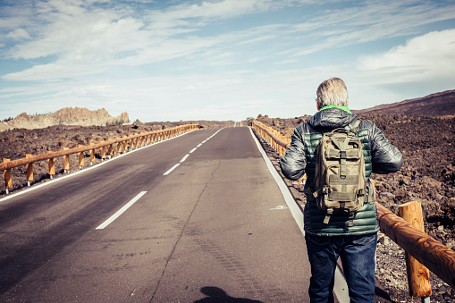 Mature retired man viewed from behind walk on a long straight asphalt road in the middle of the mountains desert with a green backpack - independence and lonely traveler concept for adult people enjoying time and life