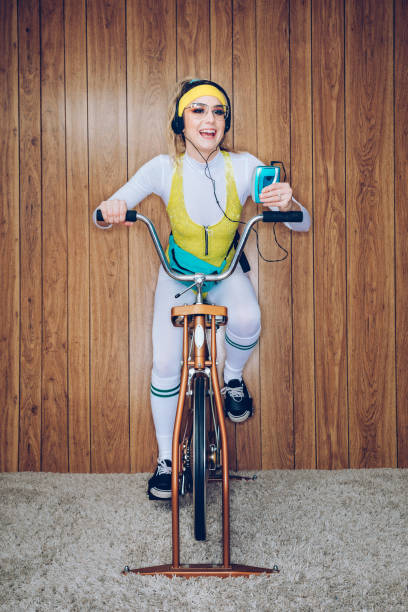 Retro Style Exercise Bike Woman Eighties Era A woman wearing exercise clothing styled after the 1980's and 1990's pedals hard on a stationary fitness bike in a vintage room, complete with shag carpet and wood paneling on the walls. She wears a leotard and a fanny pack and listens to her personal cassette tape player. walkman cassette stock pictures, royalty-free photos & images
