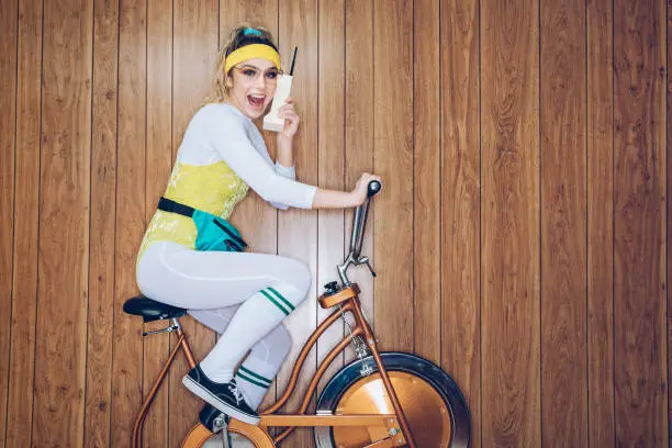 A woman wearing exercise clothing styled after the 1980's and 1990's pedals hard on a stationary fitness bike in a vintage room, complete with wood paneling on the walls. She wears a leotard and a fanny pack and talks on an early mobile phone dubbed the "brickphone".