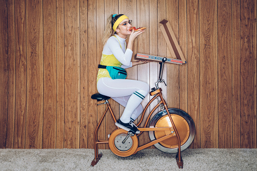 A woman wearing exercise clothing styled after the 1980's and 1990's pedals hard on a stationary fitness bike in a vintage room, complete with shag carpet and wood paneling on the walls. She wears a leotard and a fanny pack and eats from a large box of pizza.