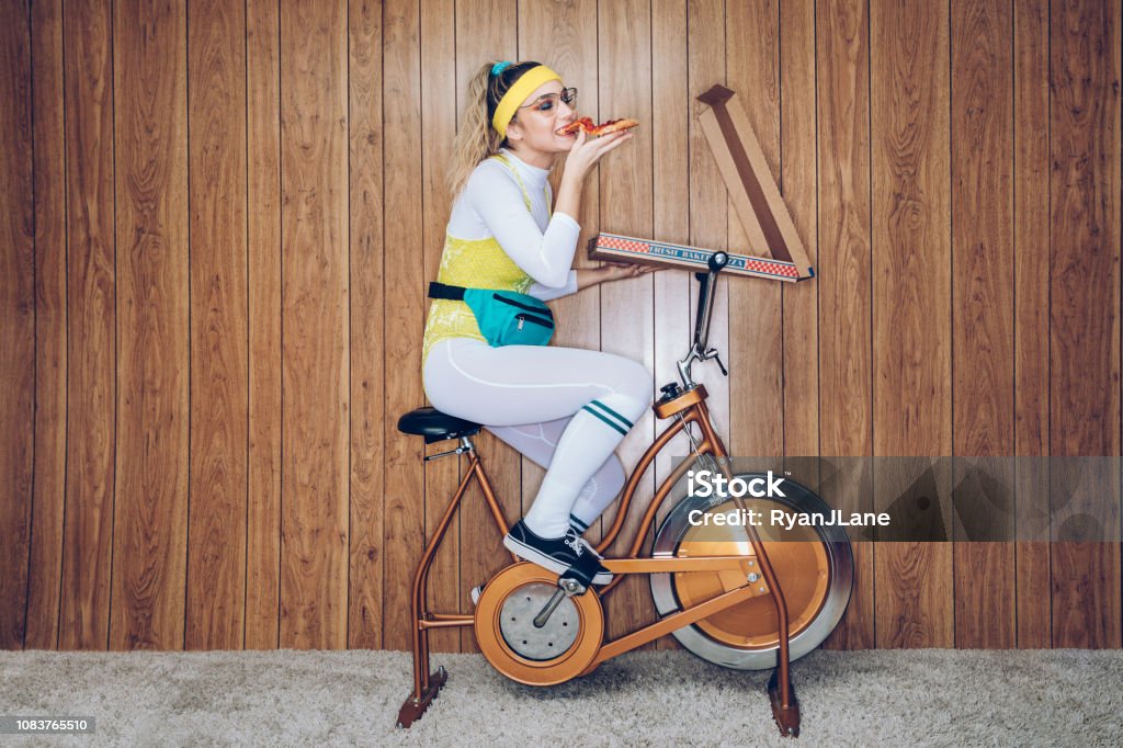 Retro Style Exercise Bike Woman Eighties Era Eating Pizza A woman wearing exercise clothing styled after the 1980's and 1990's pedals hard on a stationary fitness bike in a vintage room, complete with shag carpet and wood paneling on the walls. She wears a leotard and a fanny pack and eats from a large box of pizza. Humor Stock Photo