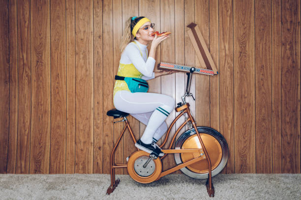 retro style exercise bike woman eighties era eating pizza - sports or fitness fotos imagens e fotografias de stock