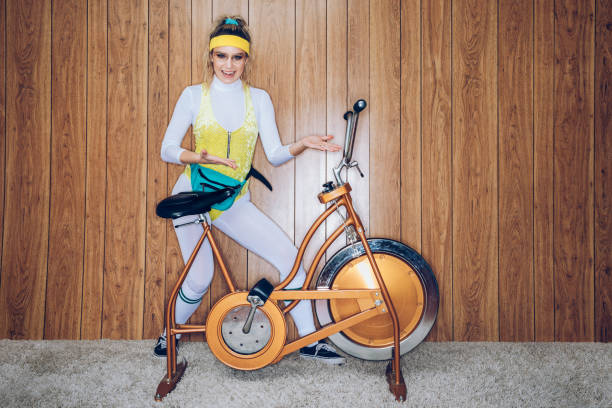 Retro Style Exercise Bike Woman Eighties Era A woman wearing exercise clothing styled after the 1980's and 1990's pedals hard on a stationary fitness bike in a vintage room, complete with shag carpet and wood paneling on the walls. She wears a leotard and a fanny pack, showing off her bike. shag rug stock pictures, royalty-free photos & images