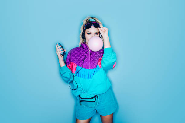 Retro Fashion Style Woman Eighties Era A woman wearing clothing styled after the 1980's and 1990's listens to music on her personal cassette tape player in front of a large bright blue background. She blows a large pink bubble with her chewing gum.  Shot with a ring flash. 1980 photos stock pictures, royalty-free photos & images