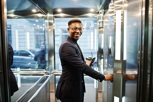 Fashionable african american man in suit and glasses with mobile phone and cup of coffee at hands posed inside elevator.