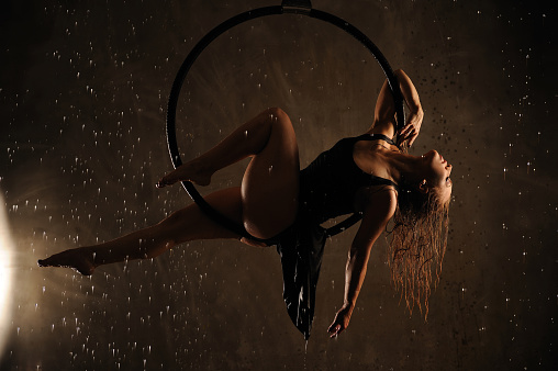 Aerial dancer during the rain. Young sports girl dancing with an air hoop. She is dressed in a black dress and high heels.