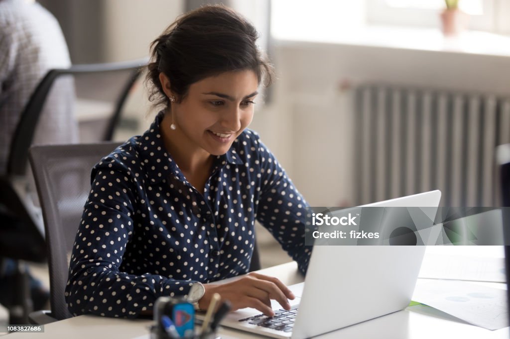 Smiling Indian female employee using laptop at workplace Smiling Indian female employee using laptop at workplace, looking at screen, focused businesswoman preparing economic report, working online project, cheerful intern doing computer work, typing Indian Ethnicity Stock Photo