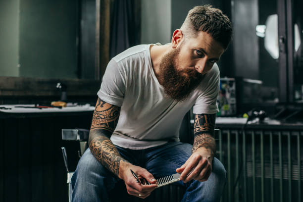 istock Calm bearded barber sitting and looking while holding comb 1083857174