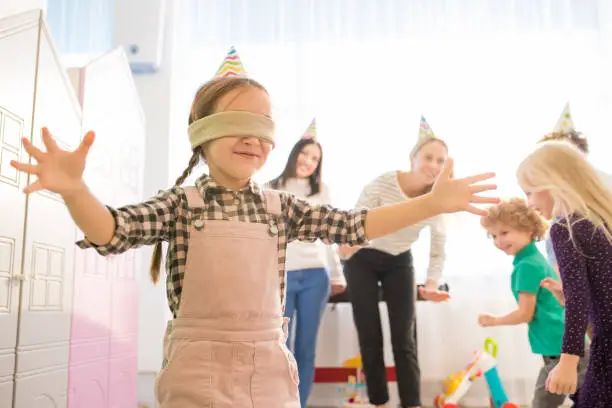 Positive content blindfolded girl in party hat keeping arms outstretched while catching up friends during blind mans buff gamer, she finding other players