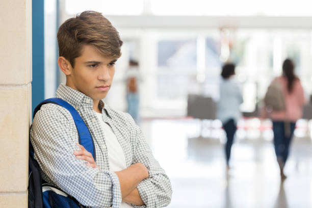 Angry male high school student stands in lobby An angry and depressed male high school student stands in his school lobby with his arms folded.  He looks down and leans against a wall with a sad expression. sad child standing stock pictures, royalty-free photos & images