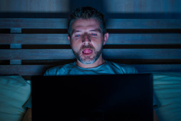 young aroused man alone in bed playing cybersex using laptop computer watching porn sex movie late at night with lascivious pervert face expression in internet pornographic sexual content stock photo