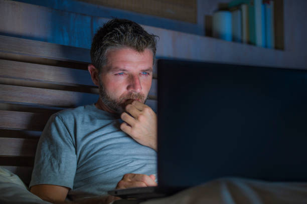 atraente e descontraído internet viciado jovem rede concentrou-se tarde da noite na cama com o computador portátil no conceito de empresário de vício ou viciado em trabalho de mídias sociais - sleaze - fotografias e filmes do acervo