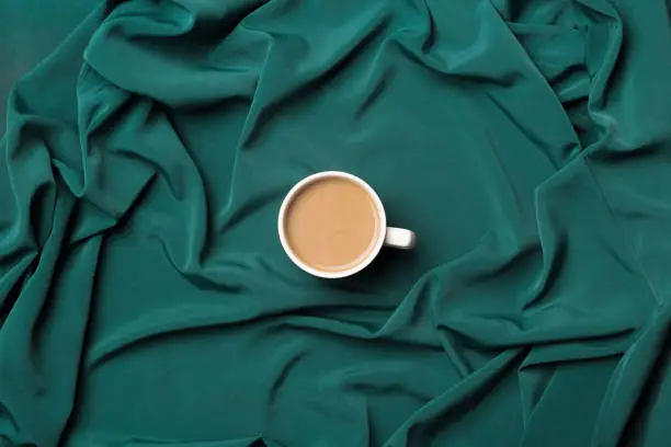 Coffee with milk in white cup on silky green fabric a background. Top view. Coffee minimal flat lay concept