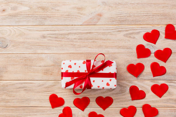 Valentine or other holiday handmade present in paper with red hearts and gifts box in holiday wrapper. Present box of gift on orange wooden table top view with copy space, empty space for design. san valentín u otro día de fiesta presente hecho a mano en papel con caja roja de corazones y regalos en la envoltura de vacaciones. presentar la caja de regalo en vista superior naranja de mesa de madera con espacio de copia, espacio vacío para el dise