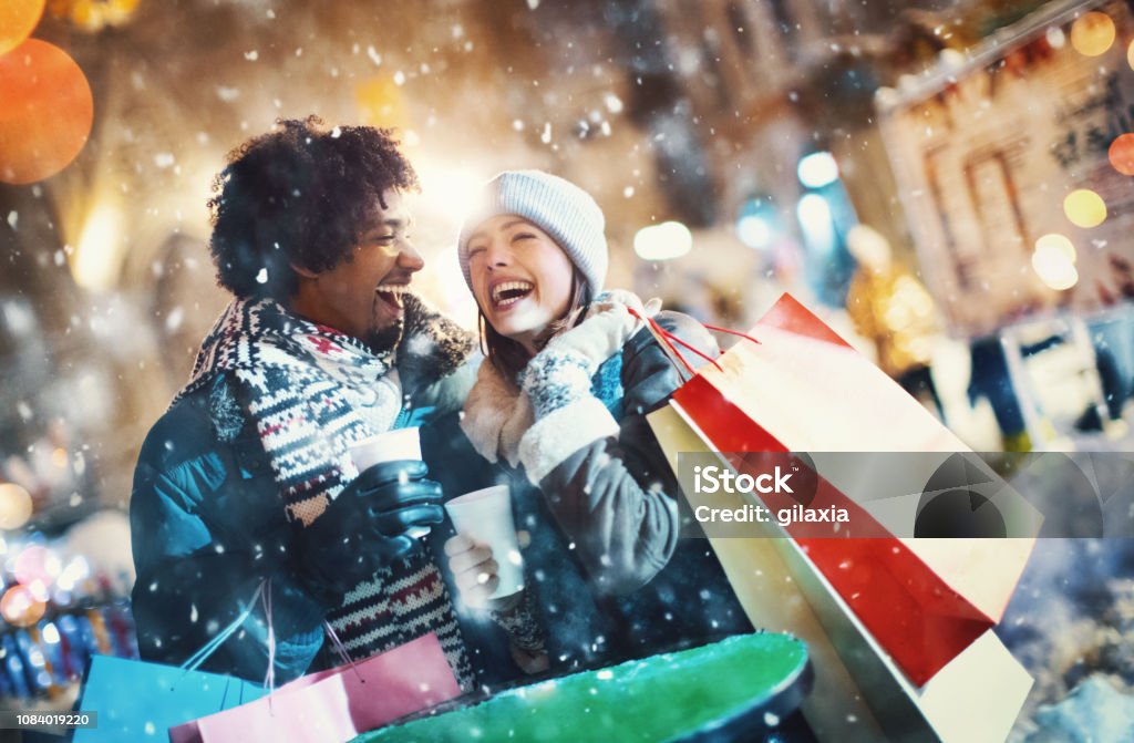 Couple doing some street shopping. Closeup front view of a happy multi ethnic couple going through Christmas market in a city square and doing some shopping. They are having fun and enjoying this cold but beautiful December night. Shopping Stock Photo