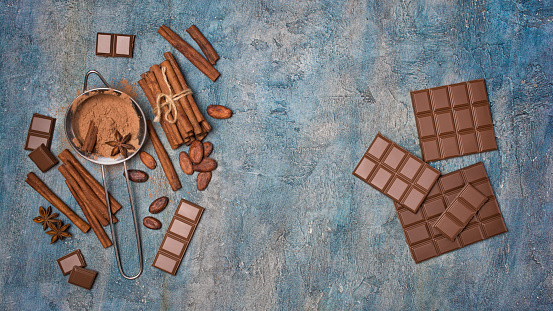 Top view of cocoa powder in strainer with chocolate, cacao beans and spices as ingredients for confectionery on blue concrete background with copy space