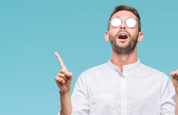 istock Young handsome man wearing glasses over isolated background amazed and surprised looking up and pointing with fingers and raised arms. 1084051704