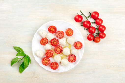 A plate of Italian Caprese salad with Mozzarella cheese, cherry tomatoes and basil leaves, shot from above on a rustic background with ingredients