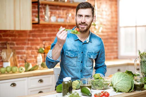 Handsome man eating healthy salad sitting at the table full of green ingredients on the kitchen at home