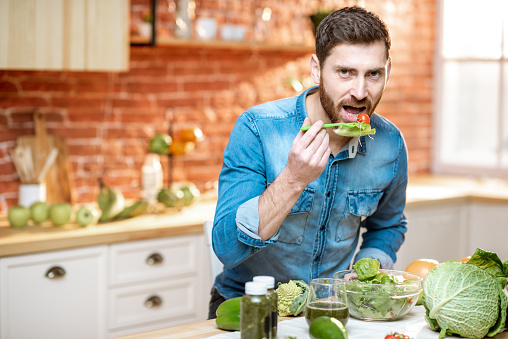 Handsome man eating healthy salad sitting at the table full of green ingredients on the kitchen at home