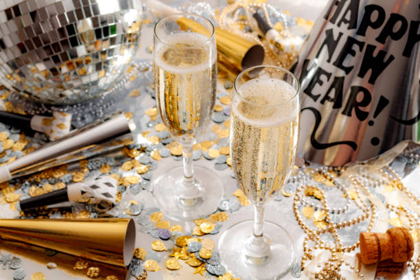 Happy new year celebration concept theme with close up on two glasses of champagne, disco ball covered in mirror, noise makers and party trumpets, confetti, beads and a cork on silver background Happy new year celebration concept theme with close up on two glasses of champagne, disco ball covered in mirror, noise makers and party trumpets, confetti, beads and a cork on silver background happy new year stock pictures, royalty-free photos & images