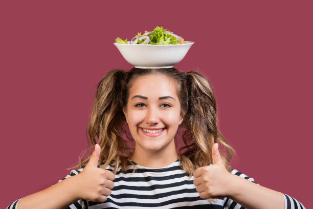 giovane ragazza felice che tiene la ciotola di insalata fresca sulla propria testa e mostra i pollici in su guardando la macchina fotografica - princess diet foto e immagini stock
