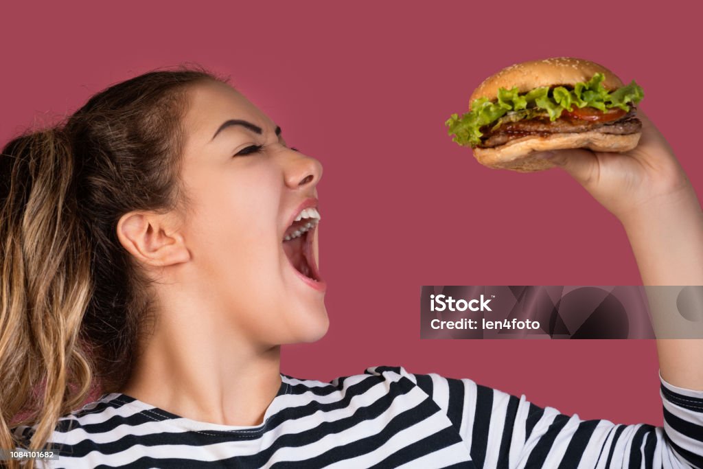 Hungry cool girl in striped t-shirt eating hamburger over pink background Crazy hungry cool girl in striped t-shirt eating hamburger in profile over colorful pink background Biting Stock Photo