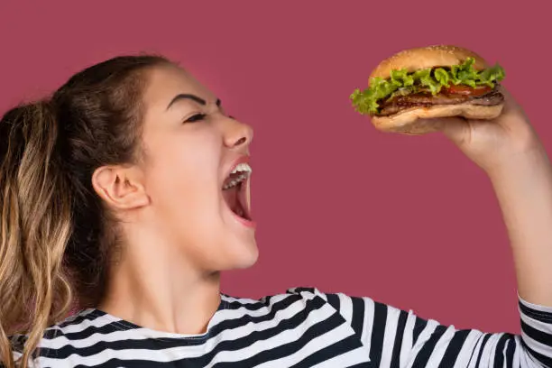 Crazy hungry cool girl in striped t-shirt eating hamburger in profile over colorful pink background