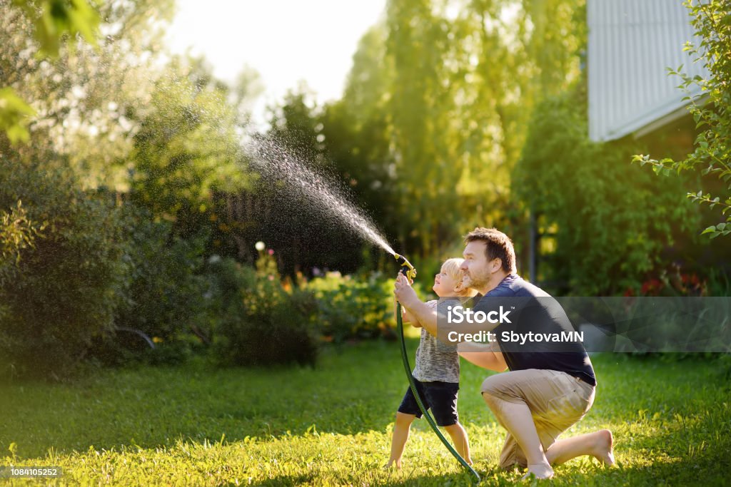 Divertido niño con su padre jugando con la manguera de jardín en patio soleado - Foto de stock de Jardín privado libre de derechos