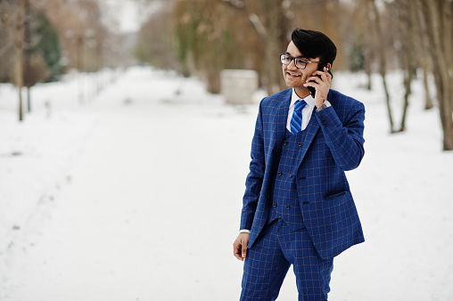 Stylish indian business man in suit and glasses posed at winter day outdoor, speaking on mobile phone.