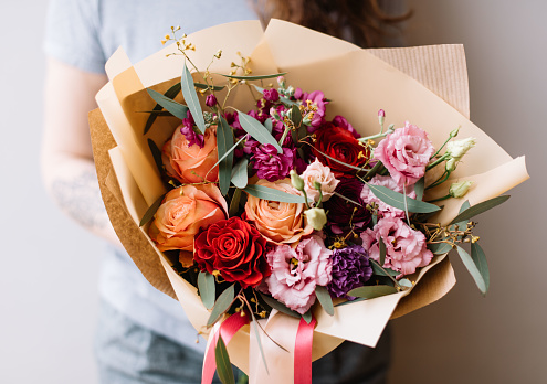 Very nice young floristvwoman holding beautiful blossoming flower bouquet of fresh roses, eustoma, carnations, eucalyptus in orange and pink colors on the grey background