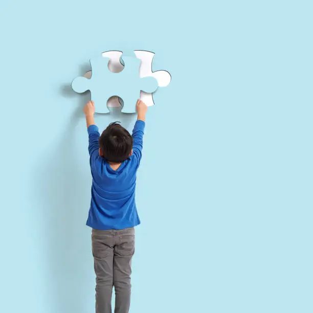 6-7 years old child holding puzzle piece on blue background. His piece complete missing piece on wall.