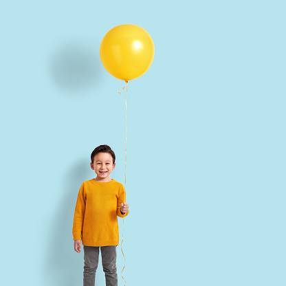 6-7 years old child holding a yellow balloon in front of blue wall.